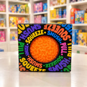 Cool Mix Squishy: Bolyhos tapintású labda 6 cm stresszlevezető játék (narancssárga), fekete, színes feliratokkal díszített dobozban, kör alakú ablakon keresztül látható.