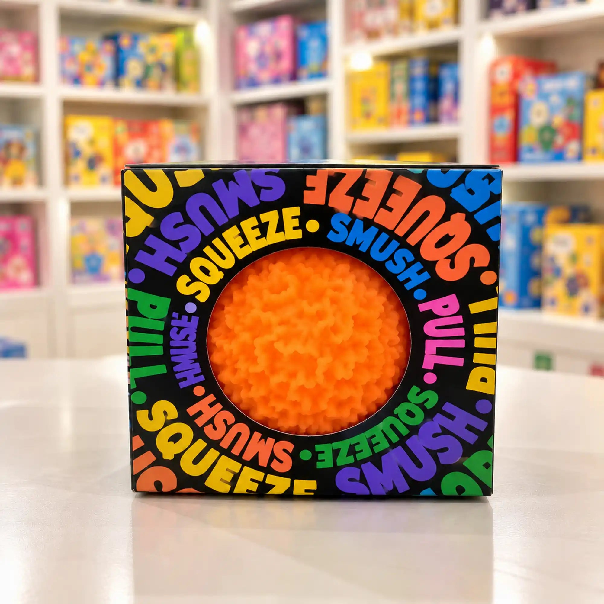 Cool Mix Squishy: Bolyhos tapintású labda 6 cm stresszlevezető játék (narancssárga), fekete, színes feliratokkal díszített dobozban, kör alakú ablakon keresztül látható.