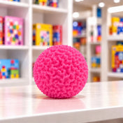 Cool Mix Squishy: Bolyhos tapintású labda 6 cm stresszlevezető játék (rózsaszín), rücskös felületű labda játékbolt pultján, színes játékdobozok előtt.
