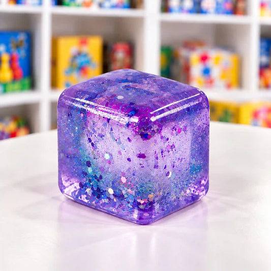 Áttetsző, lilás-kékes Cool Mix Super Gel kocka színes csillámokkal, fehér pulton, elmosódott társasjátékos polcok előtt.