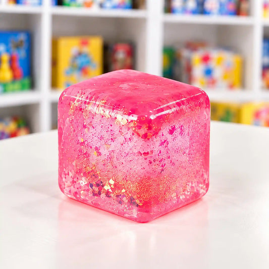 Cool Mix Squishy: Super Gel Cube stresszlevezető játék (rózsaszín), áttetsző, csillámos, lekerekített sarkú zselékocka fehér asztalon.