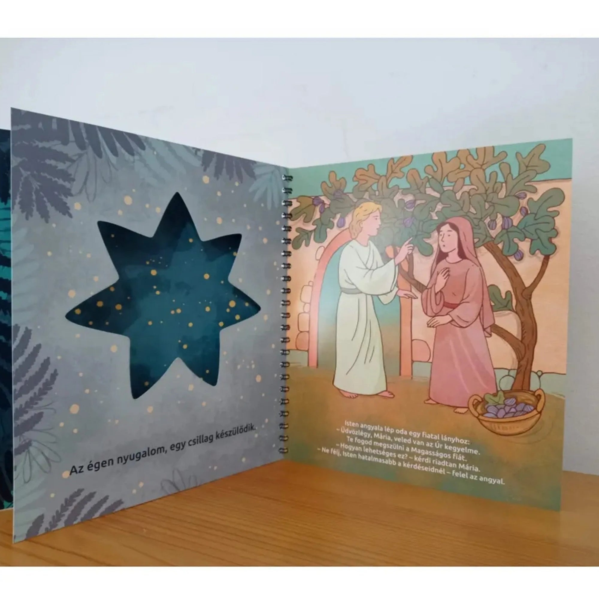 Az Ég és Föld Fia könyv oldalán csillag alakú kivágás mögött Gábriel angyal üzen Máriának a kertben.