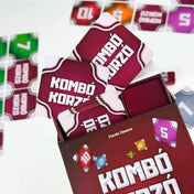 A Kombó Korzó társasjáték dobozából kilógó paklik, piros hátlapú kártyák és a háttérben színes lapok láthatók.