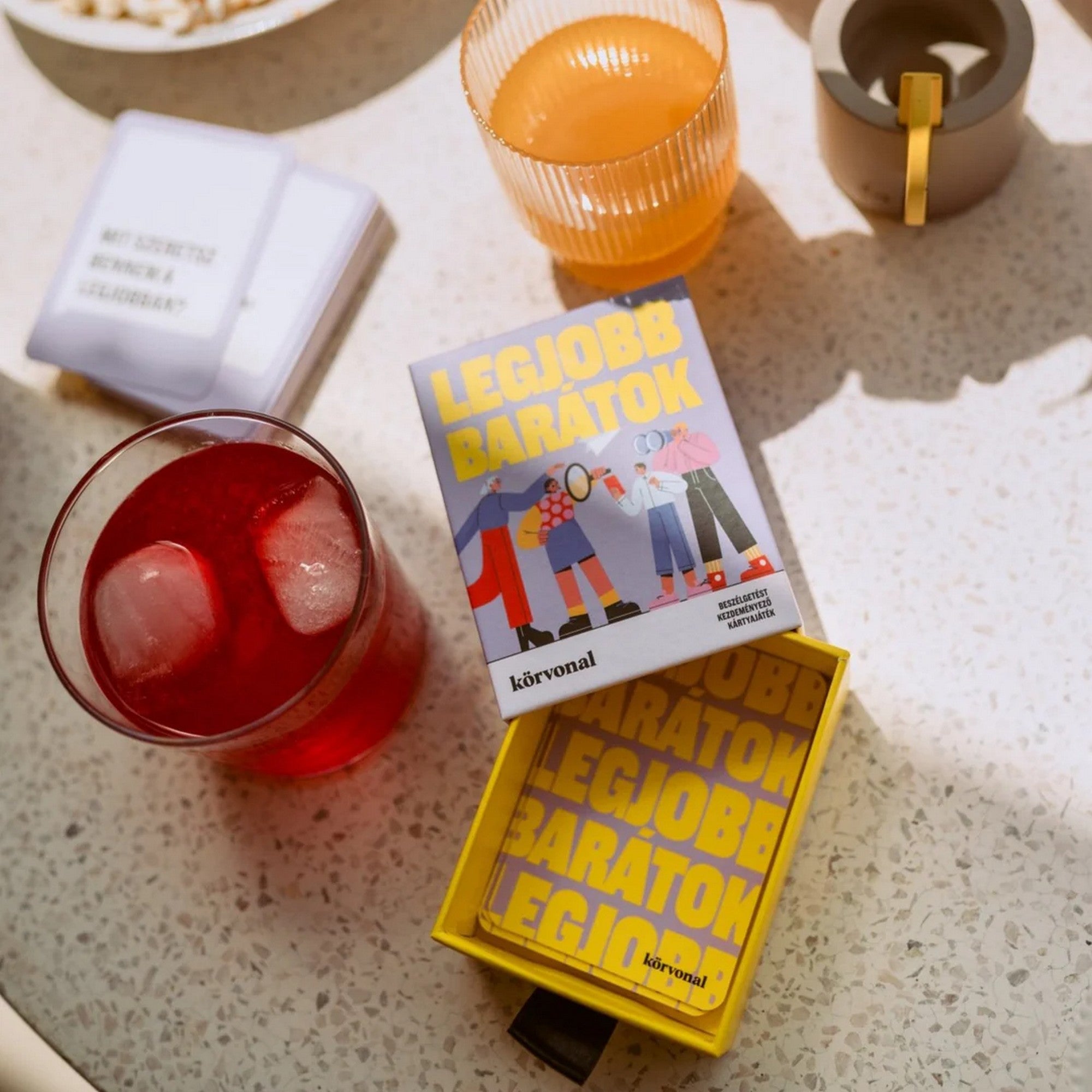 A Legjobb barátok kártyajáték doboza egy asztalon, körülötte italos poharak és néhány kártyalap. A doboz lilás színvilága és a vidám illusztráció barátságos, meghitt hangulatot sugároz.