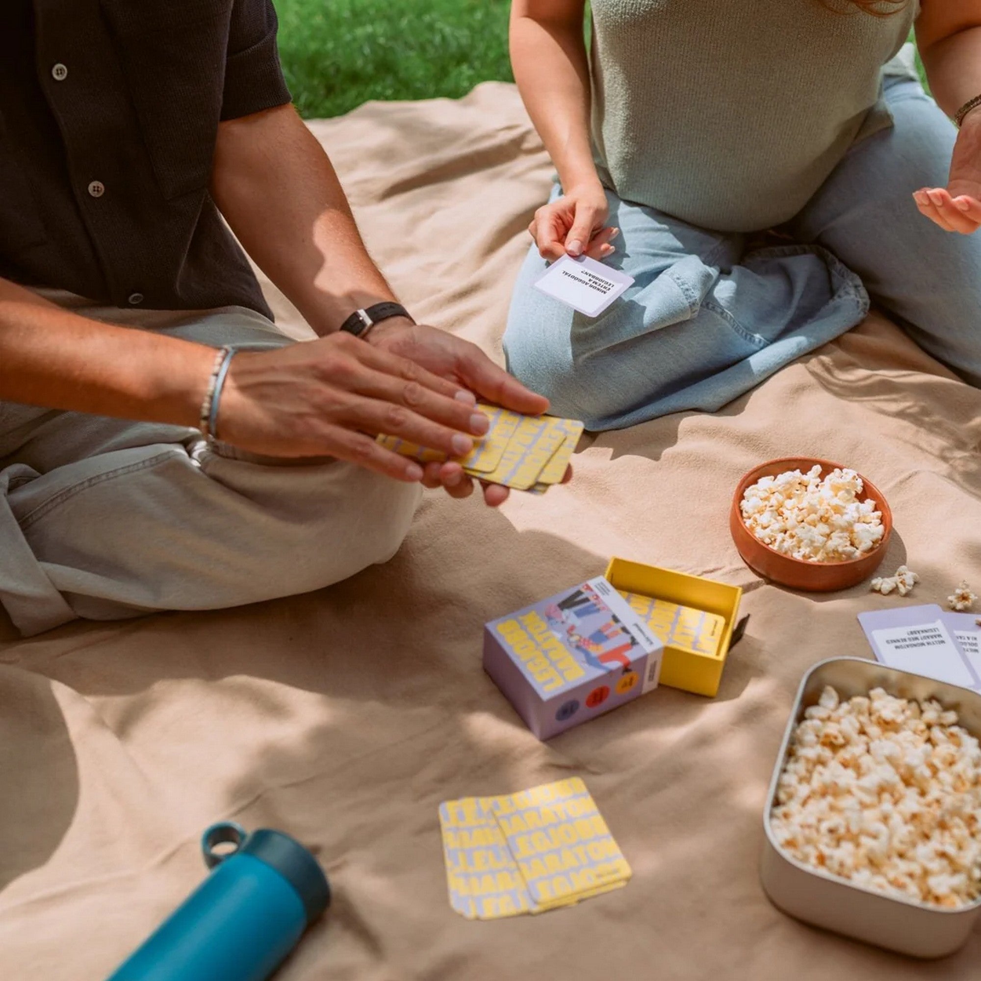 Két ember a szabadban, egy pokrócon ülve játszik a Legjobb barátok kártyajátékkal. A képen látható a nyitott doboz, sárga hátlapú kártyák és egy tál popcorn, amelyek baráti, oldott hangulatot teremtenek.