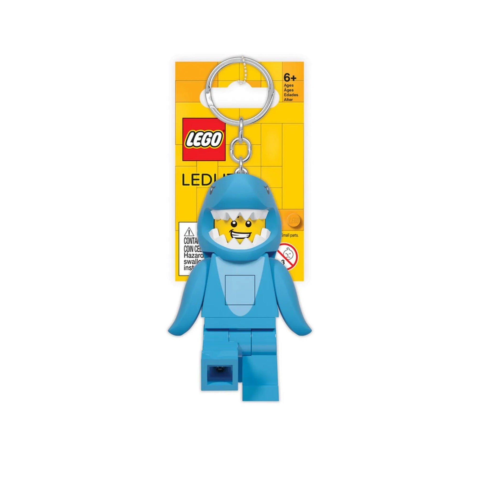 A képen a LEGO LED Shark Guy kulcstartó látható, kék cálarcú minifigura formájában, sárga LEGO háttérlapos csomagolásban. A figura vidáman mosolyog, és a kulcstartó tetején ezüstszínű fémkarika található.