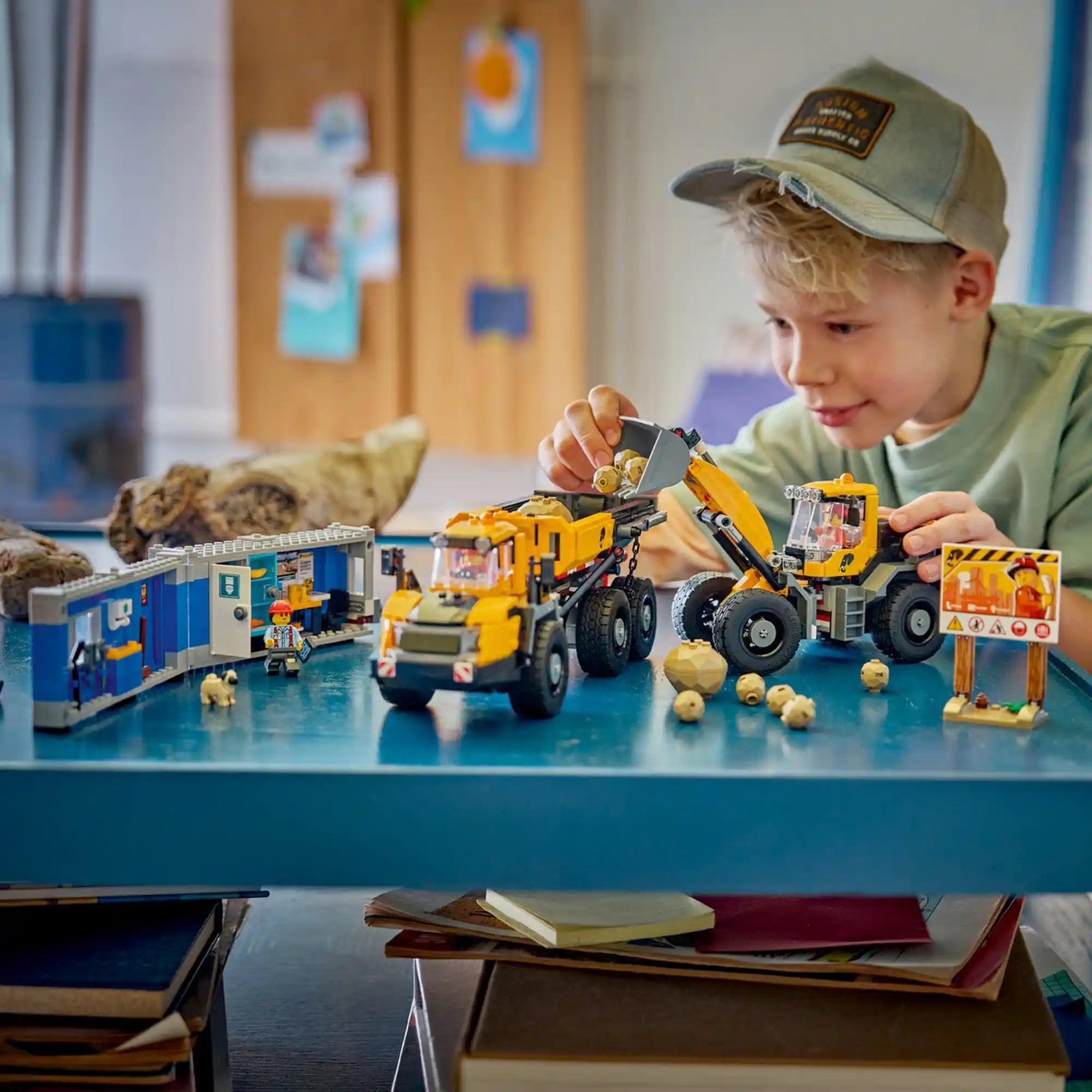 Kisfiú játszik a LEGO City 60494 építési munkagépekkel, miközben a homlokrakodó köveket rak a dömperbe, a háttérben pedig a mobil iroda és az építkezési tábla látható.