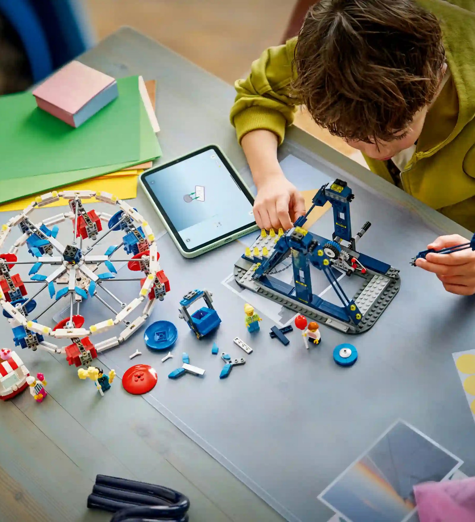 Gyermek építi a LEGO Creator 3 az 1-ben Ikonikus óriáskerék (31389) készletet. Az asztalon részben összeépített óriáskerék, szétszórt LEGO elemek, minifigurák és a LEGO Builder alkalmazást futtató táblagép látható.