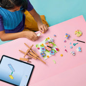 Gyermek építi a LEGO Creator 3 az 1-ben Virágos művészeti alkotások (31390) készletet. Az asztalon szétszórt színes elemek, a virágos műalkotás alapja, festőállvány alkatrészei és a LEGO Builder alkalmazást futtató táblagép látható.