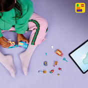 Egy kisgyermek ül a földön, és a LEGO® ǀ Disney Ariel varázslatos minipalotája (43285) készlettel játszik. A kép bal oldalán látható az épülő minipalota, mellette szétszórt LEGO elemek, figurák (Ariel, Sebastian, Ficánka), kincsesláda és egy tablet, amin a LEGO Builder alkalmazás fut, lila háttérrel.