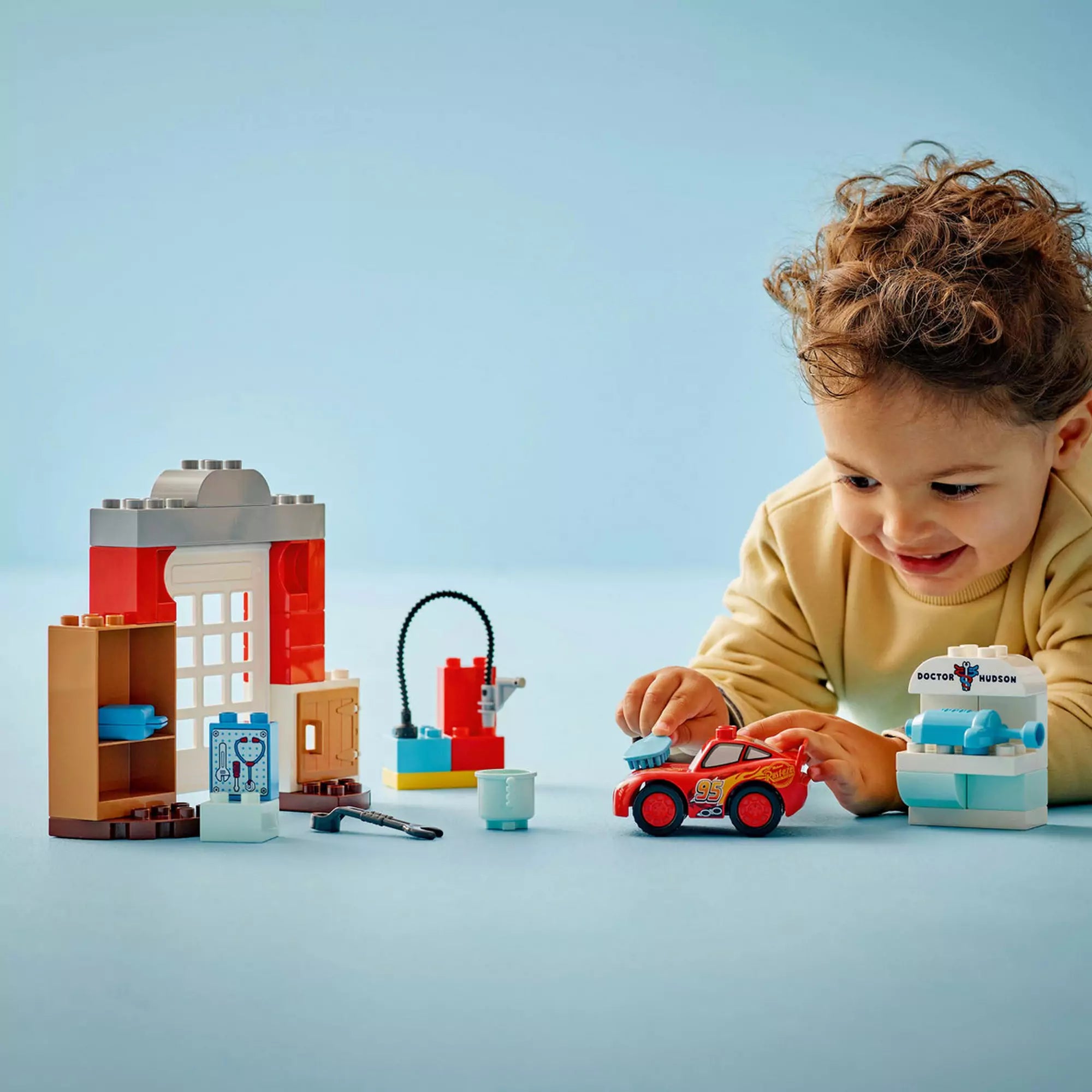 Kisgyermek mosolyogva játszik a LEGO DUPLO McQueen látogatása Doc garázsában (10456) készlettel, miközben Villám McQueen-t egy vizsgálóeszközzel kezeli.