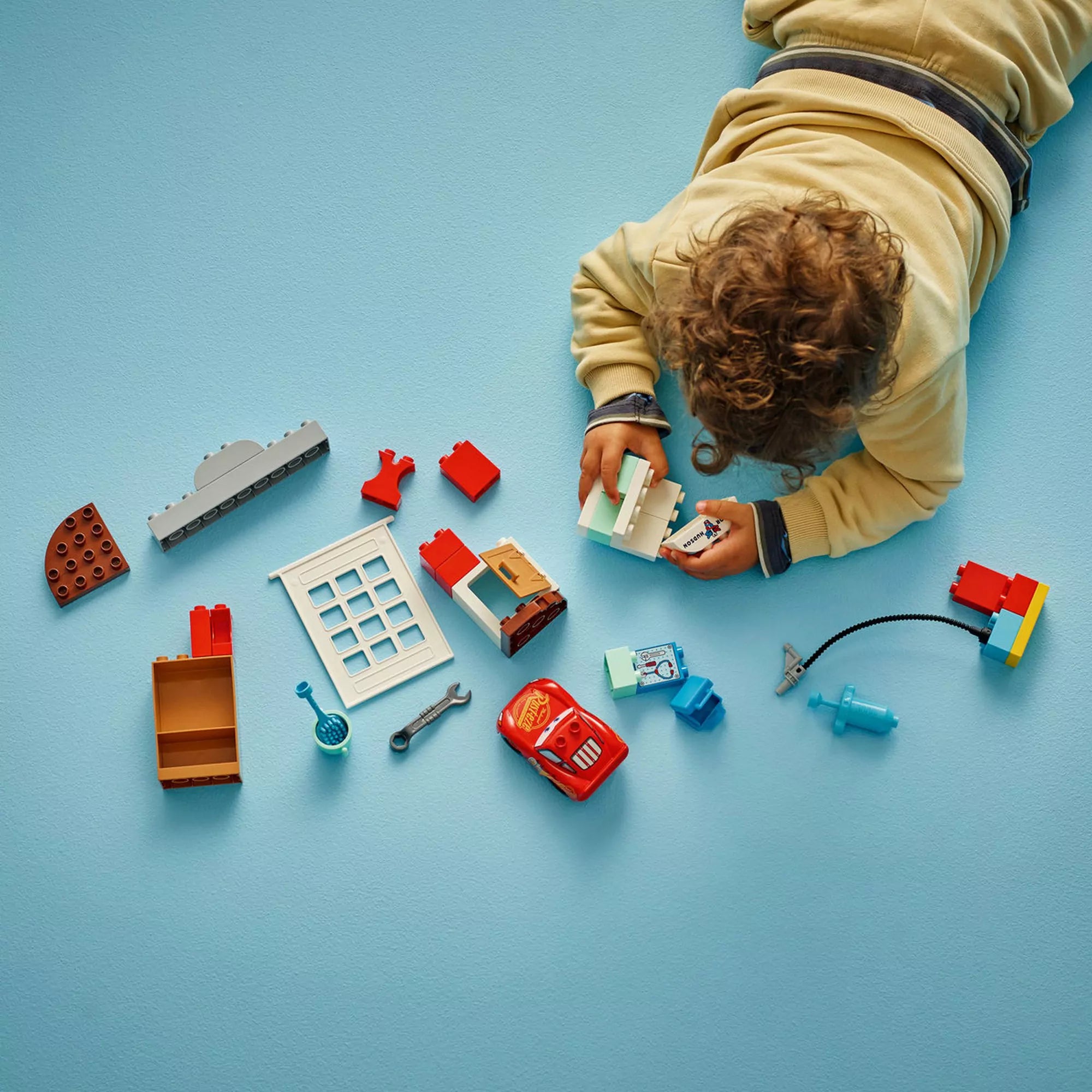Kisgyermek fentről fotózva, épp a LEGO DUPLO McQueen látogatása Doc garázsában (10456) elemeit szereli a világoskék padlón.