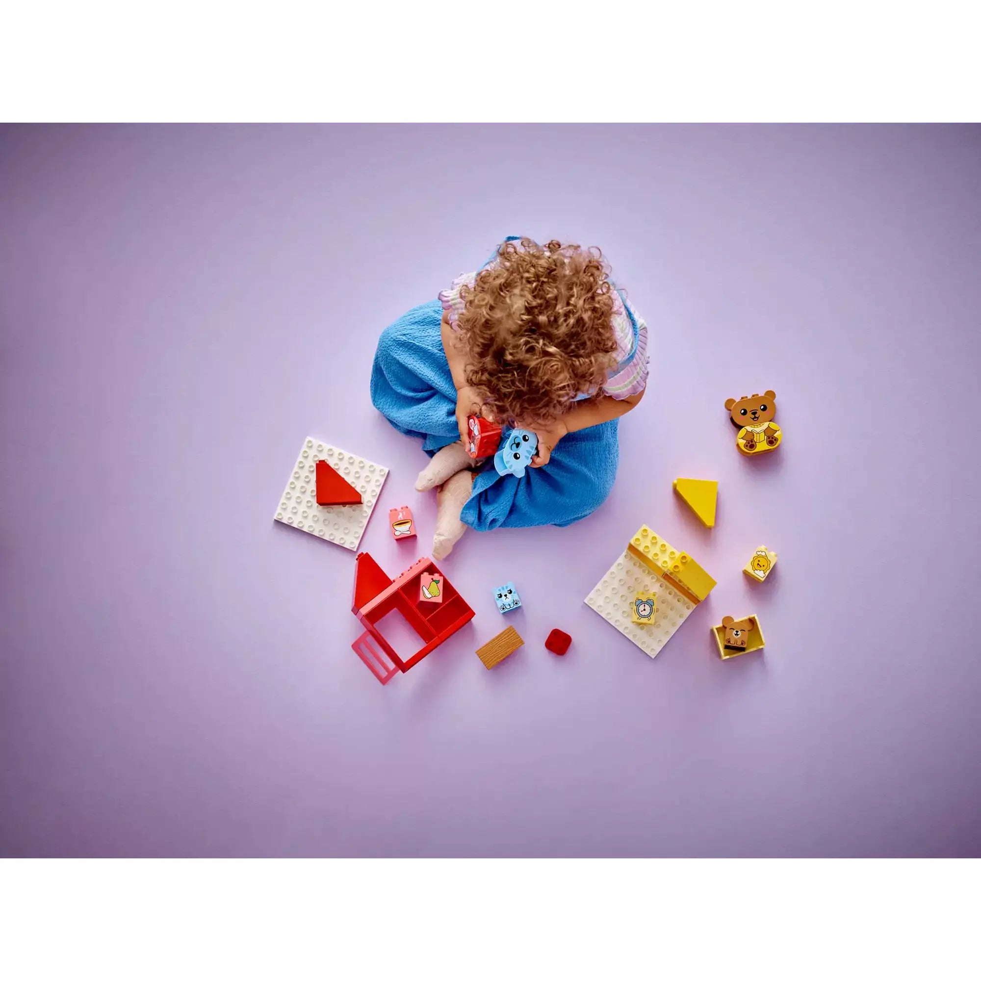 LEGO Duplo játék madártávlatból, szétszedve – Egy kislány ül a padlón és körülötte a LEGO Duplo 10414 készlet elemei, köztük háztetők, figurák és ételmotívumok.