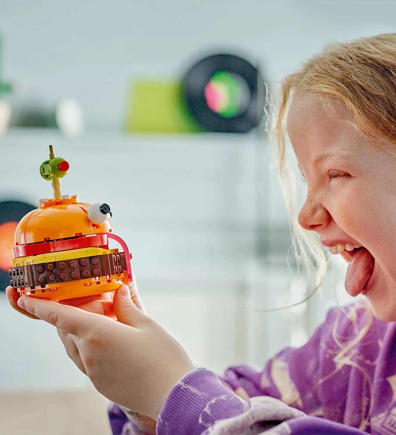 Egy mosolygó kislány kezében tartja a megépített LEGO Fortnite Durrr Burger modellt, miközben nevetve utánozza a figura kinyújtott nyelvét. A kép jól bemutatja a játék vidám hangulatát.