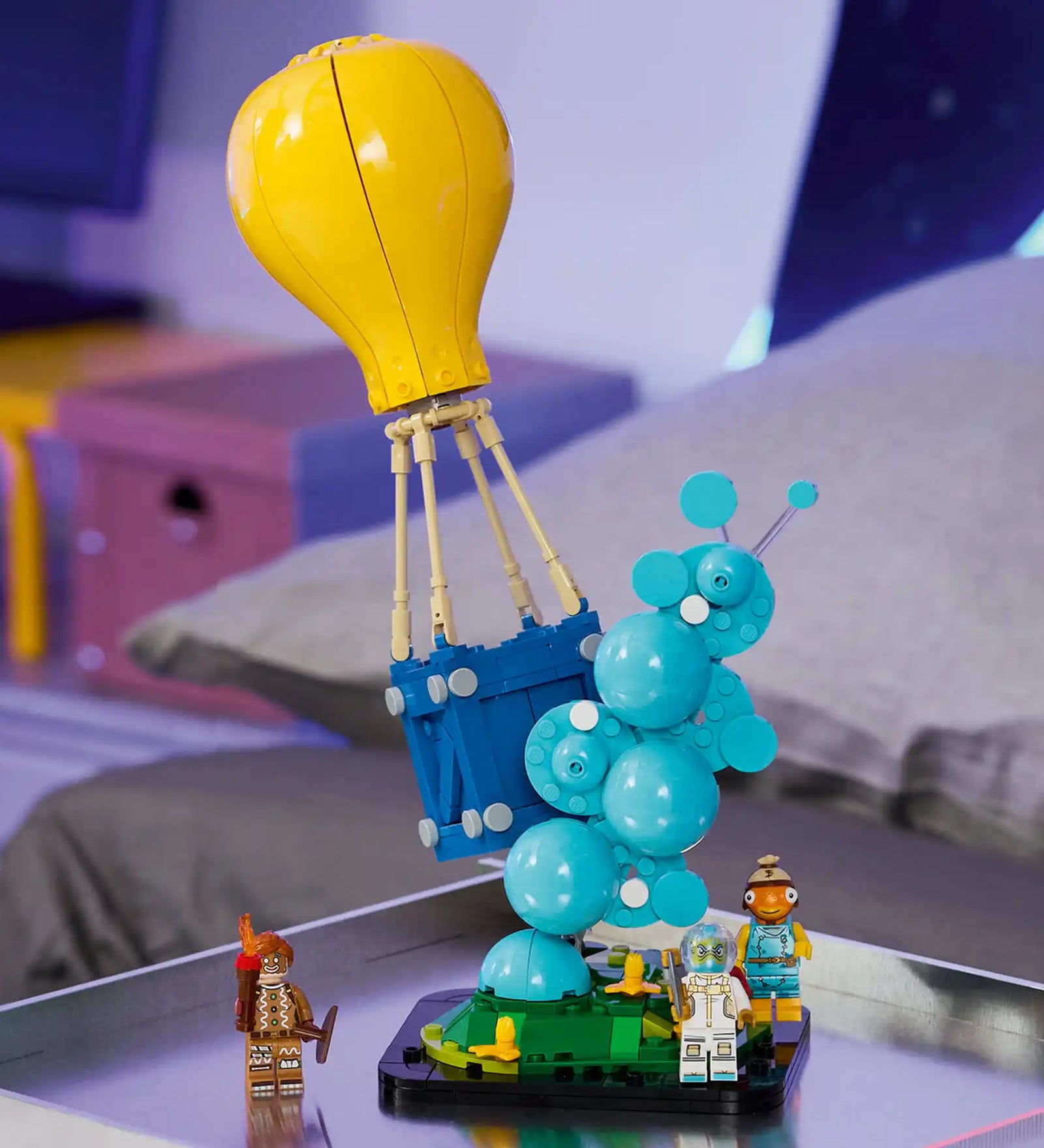 Megépített LEGO Fortnite Supply Drop modell kiállítva, ikonikus sárga léggömbbel, kék ellátmányládával és három Fortnite karakterrel.