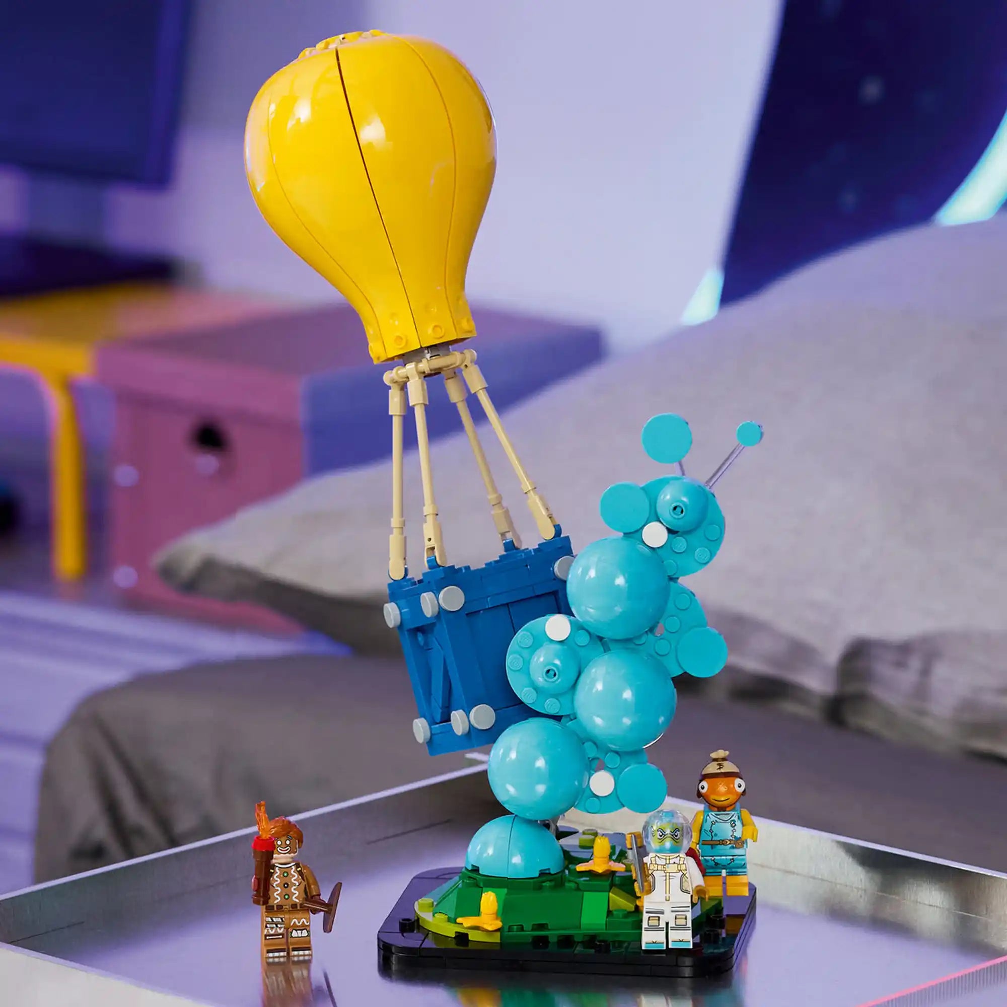 Megépített LEGO Fortnite Supply Drop modell kiállítva, ikonikus sárga léggömbbel, kék ellátmányládával és három Fortnite karakterrel.