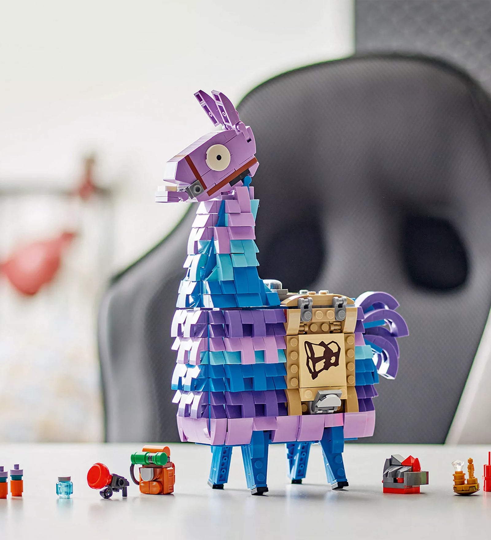 A megépített LEGO Fortnite Supply Llama figura asztalon kiállítva, a játékban ismert ikonikus színekben: lila, kék és türkiz. A láma mellett apró építhető Fortnite kiegészítők láthatók, például hátizsák, italok és dinamitrúd.