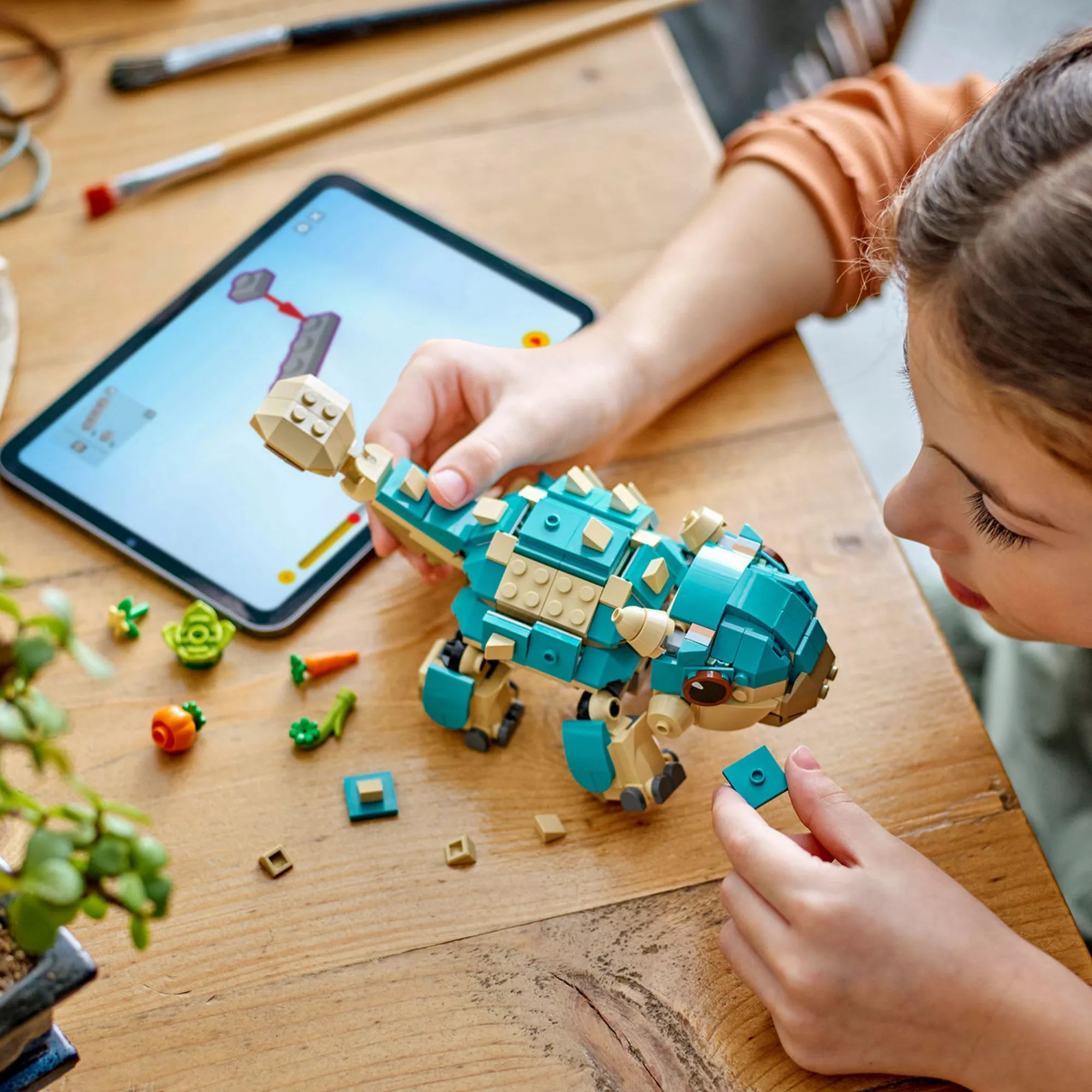 „Egy gyermek építi a LEGO Bumpy bébidínó figurát az asztalnál, mellette a zöldségkiegészítők és a LEGO Builder alkalmazás fut a táblagépen.”