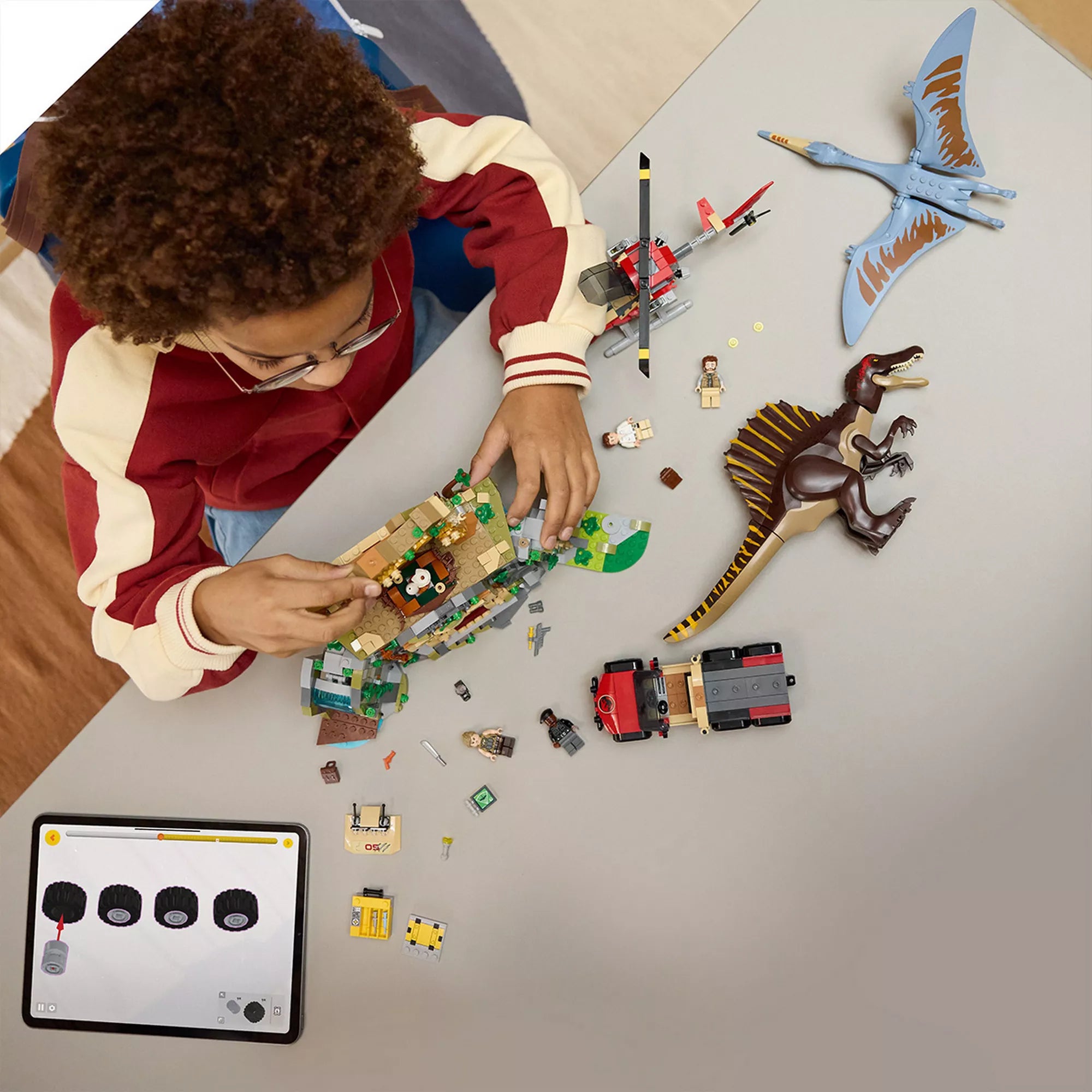 „A LEGO Spinosaurus és Quetzalcoatlus légi küldetés (76976) elemei szétszórva az asztalon, köztük dinoszaurusz figurák, templom, helikopter, terepjáró és minifigurák, építés közben táblagépes útmutatóval.”