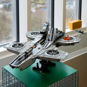 A szürke LEGO Helicarrier fekete állványon, zöld asztalon, ablak előtt kiállítva, a fedélzeten lángokkal és Quinjet repülővel.