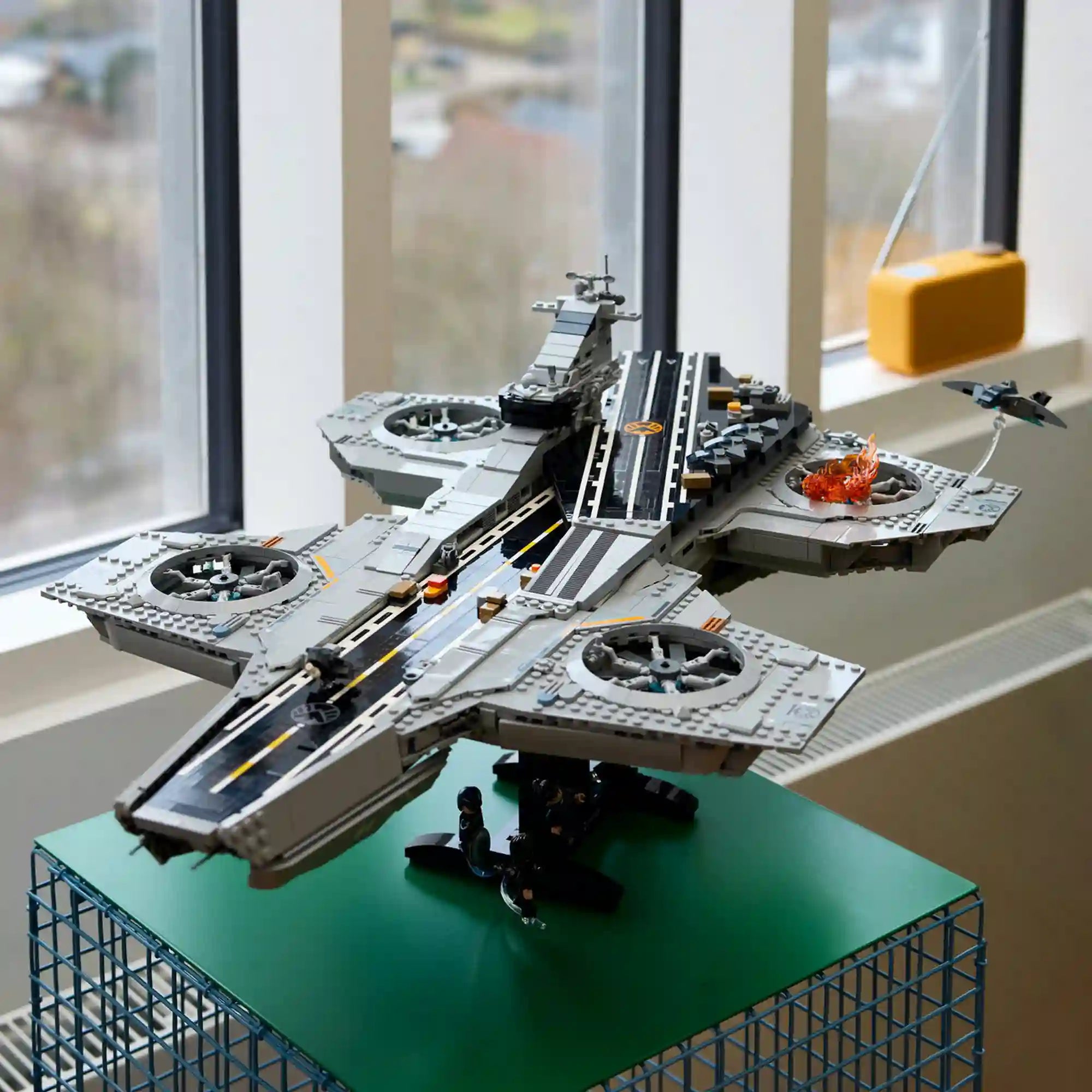 A szürke LEGO Helicarrier fekete állványon, zöld asztalon, ablak előtt kiállítva, a fedélzeten lángokkal és Quinjet repülővel.