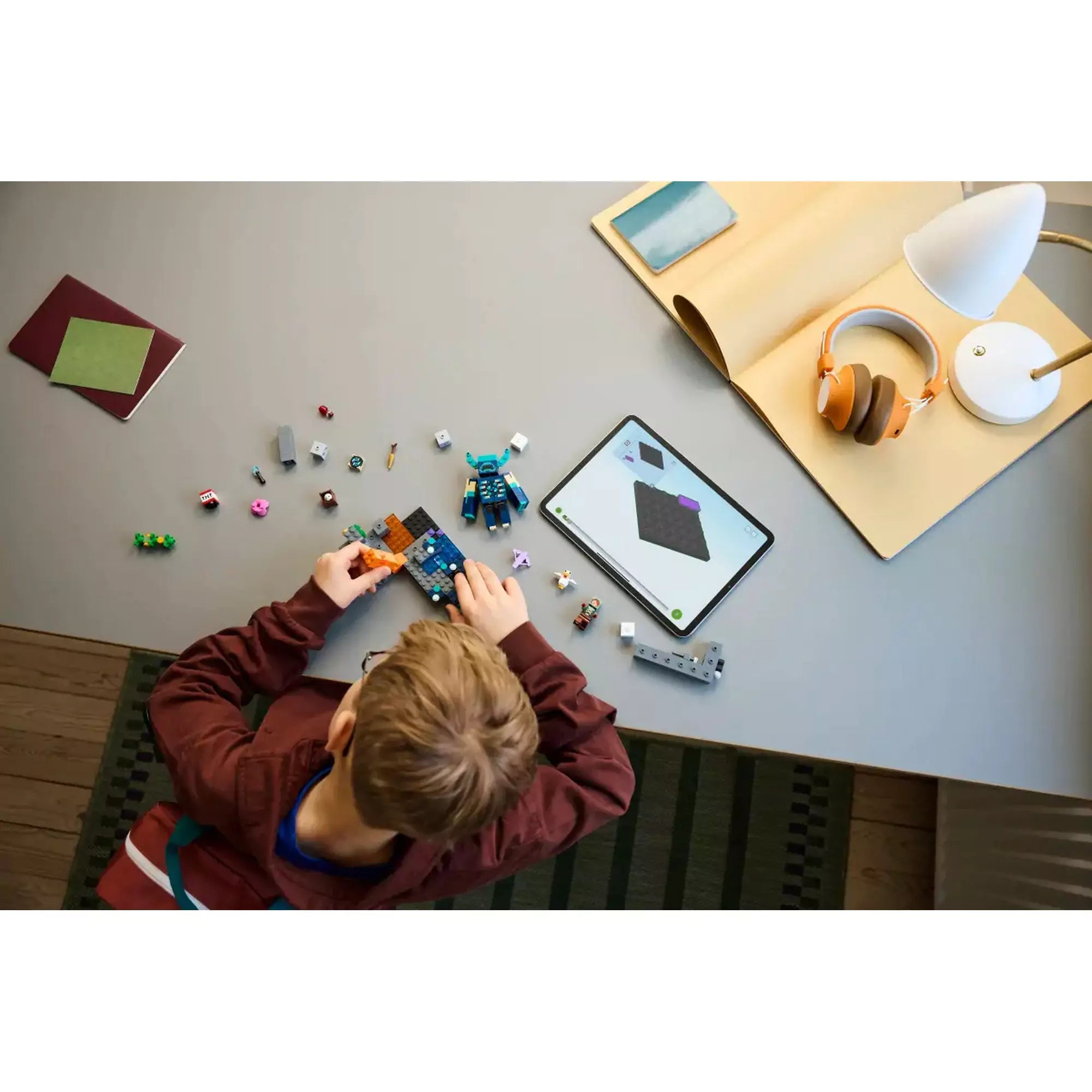 Egy gyerek építi a LEGO Minecraft Találkozás az Őrzővel készletet egy asztalon, mellette tablet az interaktív digitális útmutatóval.