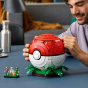 Egy férfi a LEGO Pokémon: Ikonikus Trénerpillanatok Pokélabda 72154 piros-fehér modelljét igazítja, mellette Pokémon-figurák láthatók.