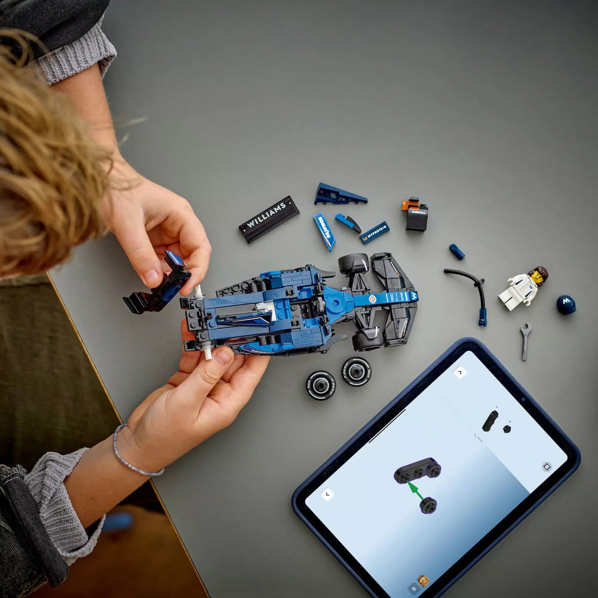 Gyermek építi a LEGO Speed Champions Williams FW46 autót, mellette tablet mutatja a digitális útmutatót és a szétszedett elemeket.