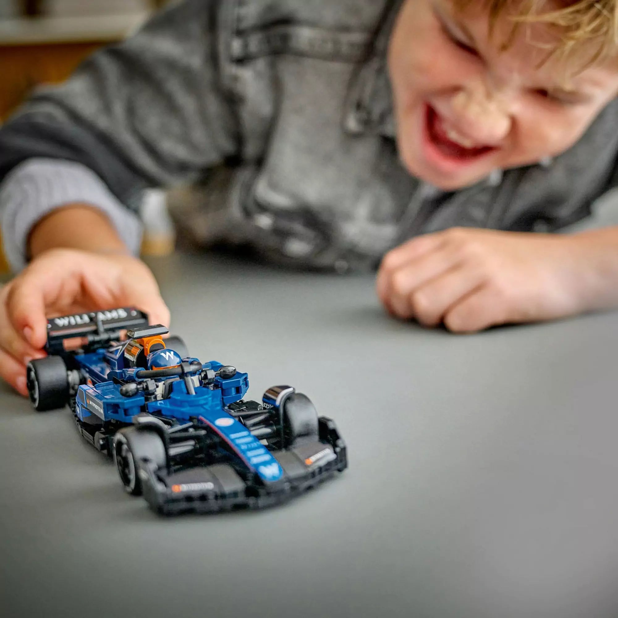 Vidám kisfiú játék közben a LEGO Speed Champions Williams FW46 versenyautóval, amelyet az asztalon tologat.