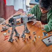 Fiatal férfi a nyitott oldalú LEGO Star Wars AT-AT lépegetőt szereli egy faasztalon, LEGO elemekkel, figurákkal és tablettel körülvéve.