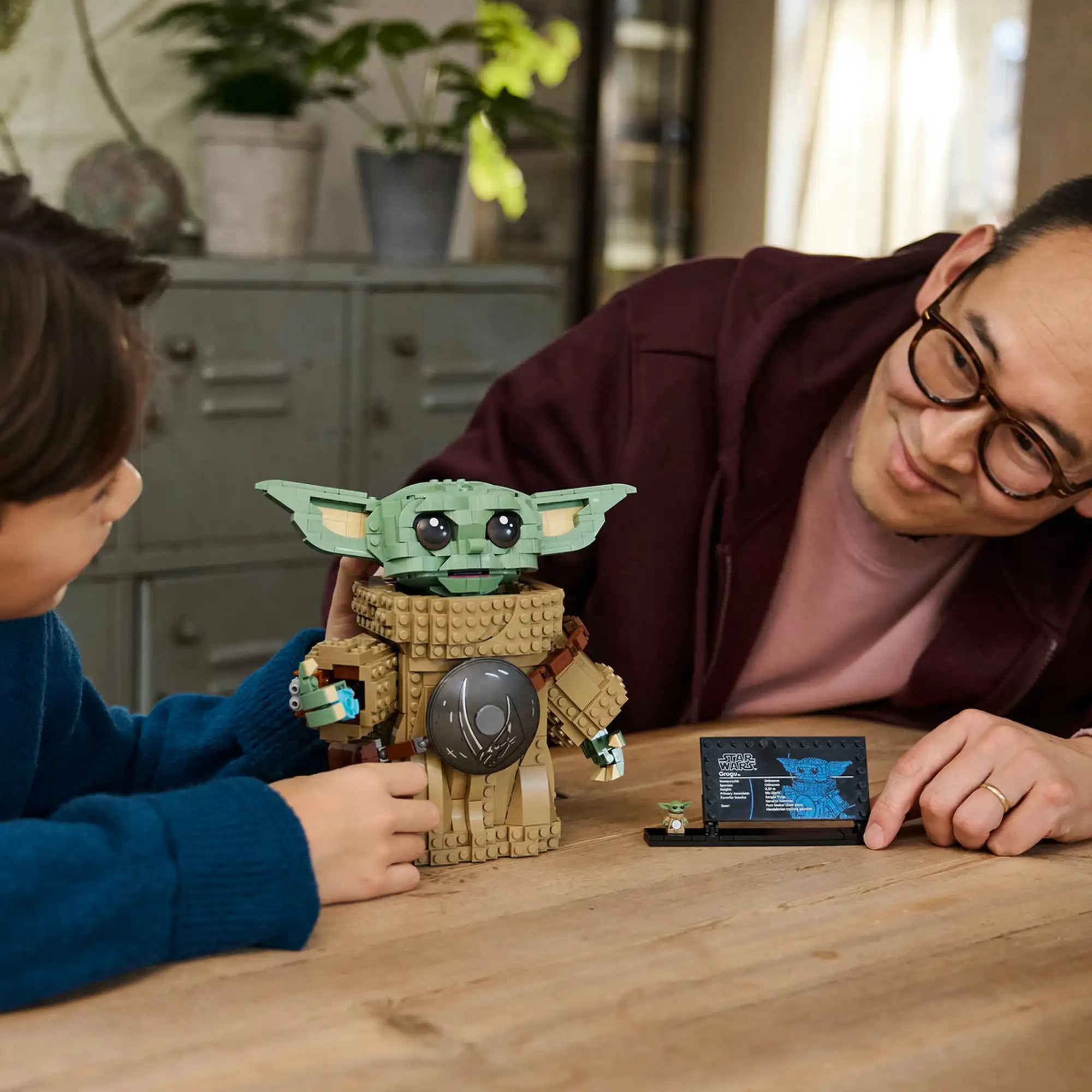LEGO Star Wars: Grogu (Mandalóri tanonc) megépített figura gyerek és felnőtt kezében, játék közbeni jelenet asztalon.