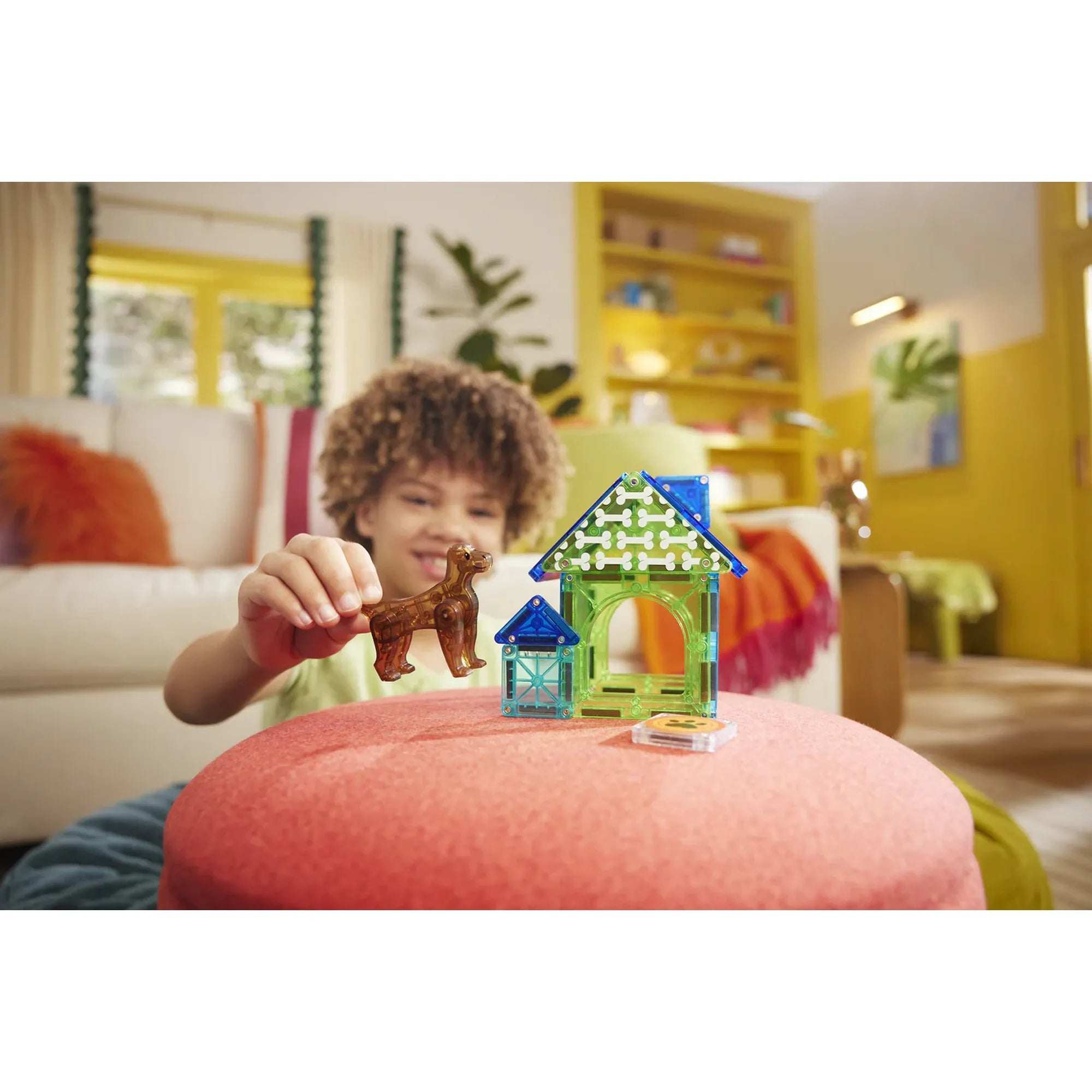Mosolygó gyermek játszik a Magna-Tiles Dog House szettel, a kezében a barna kutya figura, előtte a színes kutyaház.