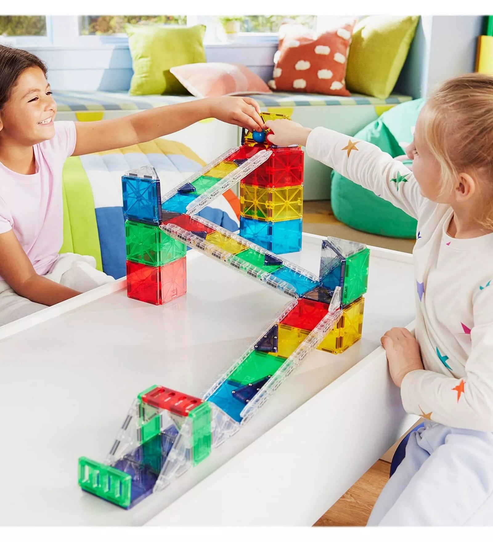 Két mosolygó gyerek épít és játszik a MAGNA-TILES Rail Racers Deluxe mágneses pályával. A színes elemekből készült pályán golyókat indítanak el, miközben közösen alkotnak és tanulnak.