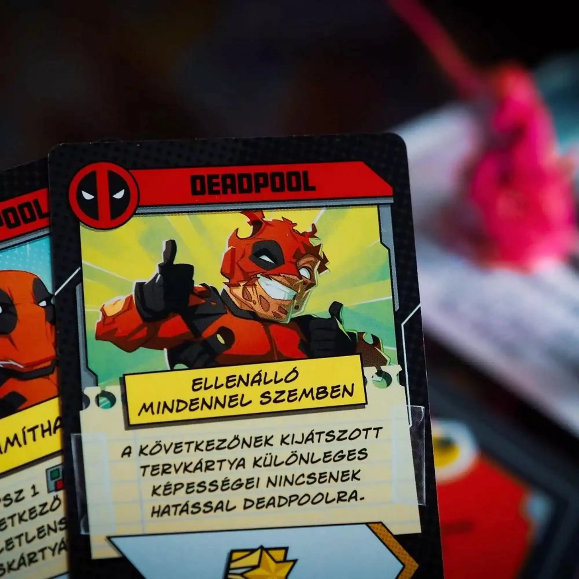 Közelkép egy Deadpool karakterkártyáról, amelyen Deadpool sérült maszkkal mosolyog és felfelé mutató hüvelykujjal pózol. A háttérben életlenül egy figura látható.
