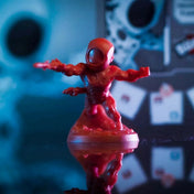 Scarlet Spider vörös miniatűr figurája akciópózban, fényes felületen, háttérben elmosódott játékelemekkel.