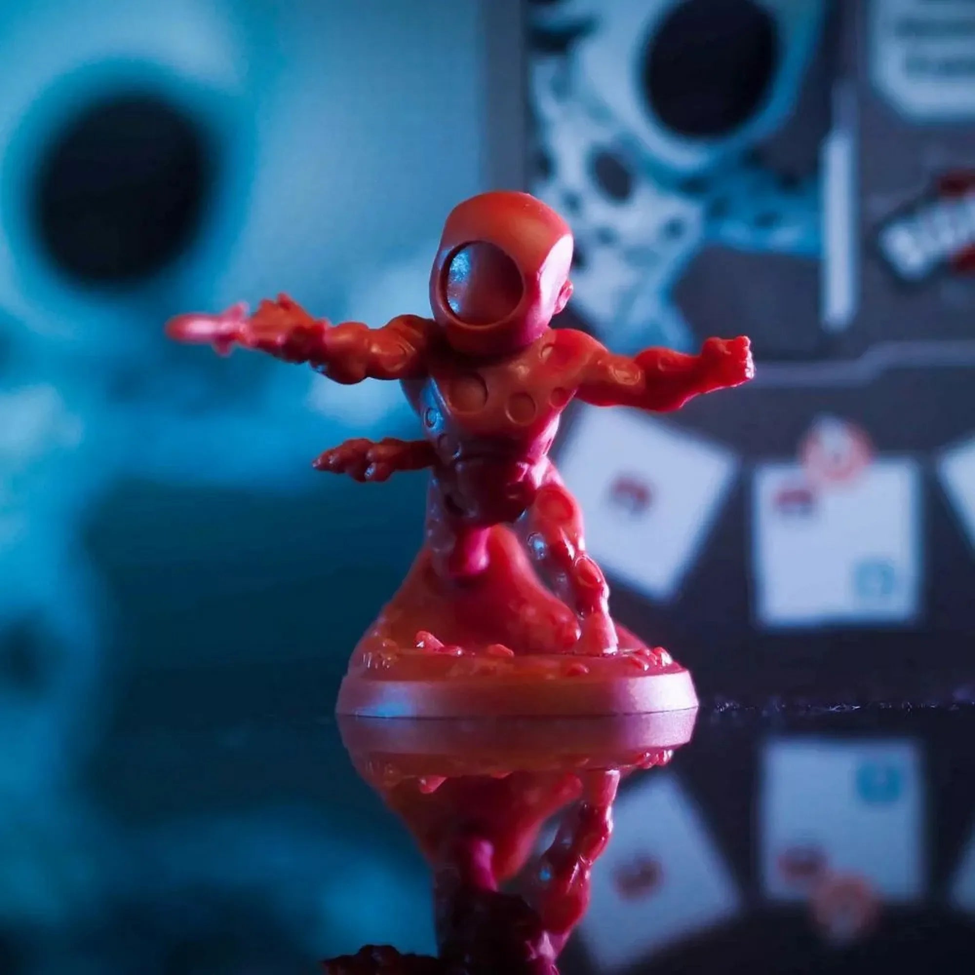 Scarlet Spider vörös miniatűr figurája akciópózban, fényes felületen, háttérben elmosódott játékelemekkel.