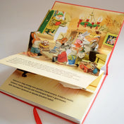 Mesél az erdő – Karácsony nyitott oldal – A könyv felnyíló jelenetében a nyuszik vidáman karácsonyoznak, az asztalon sütemény és gyertyafény teremt meghitt hangulatot.