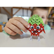 Egy gyermek kezében tartja a 3D Origami – Eper figura (Alexander) szettből készült vidám, piros-zöld színű papírfigurát, amely éppen elkészült az asztalon.