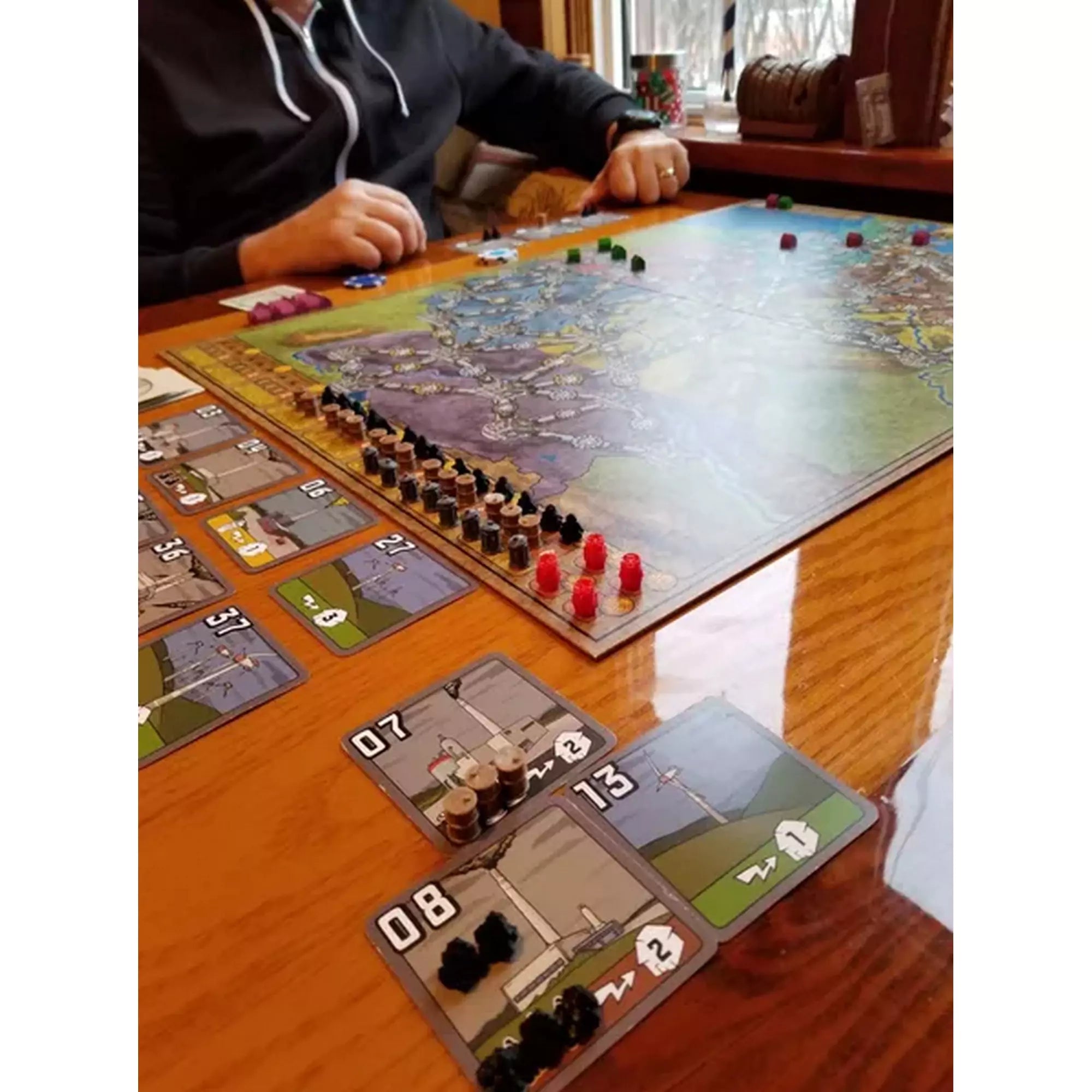 Egy asztalon elhelyezett játéktábla, kártyák és erőforrásjelölők, valamint egy játékos, aki éppen a Power Grid társasjátékkal játszik.