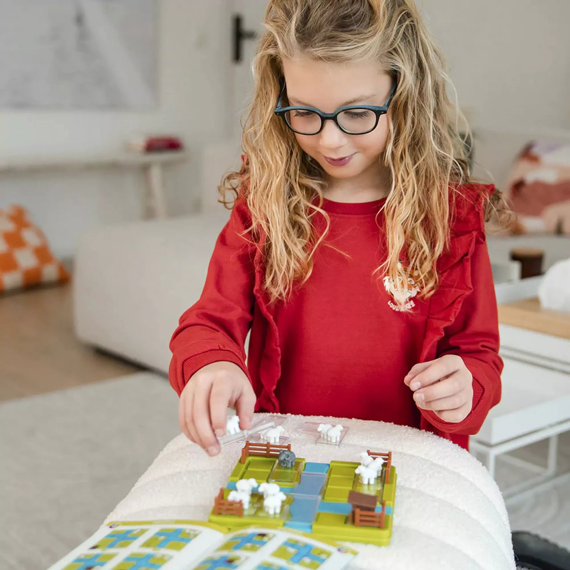 Kislány piros pulóverben játszik a Smart Games Counting Sheep logikai játékkal, figyelmesen elhelyezve a bárányokat.