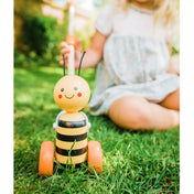 A tologatható fa méhecske játék fűben, egy kisgyermek mellett. A vidám arcú, csíkos méhecske narancssárga kerekeken áll, miközben a gyermek a tolórudat fogja. A kép játékos, kültéri jelenetet ábrázol.